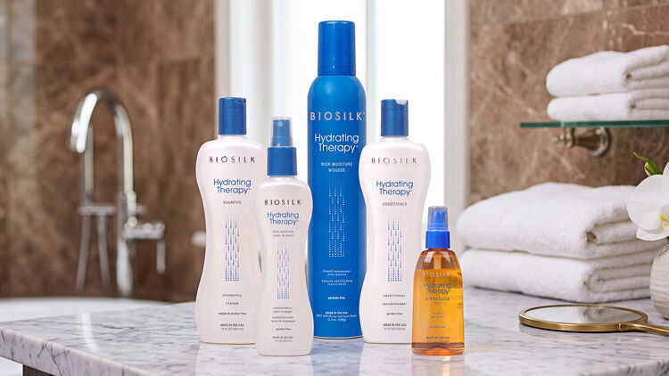 Five Biosilk Hydrating Therapy products with blue caps on a marble counter with white towels and an orchid.