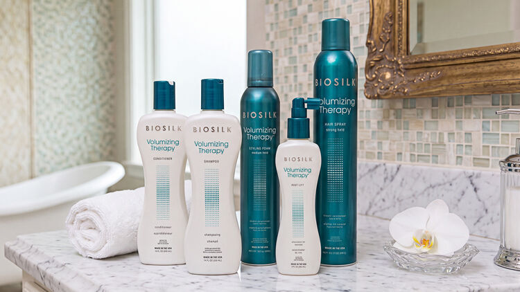 Five Biosilk Volumizing Therapy hair products with teal caps on a marble bathroom counter with a towel and orchid.