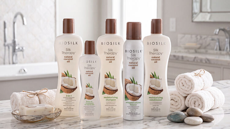 Five Biosilk Silk Therapy coconut oil hair products with brown caps on a marble counter with rolled towels and stones.